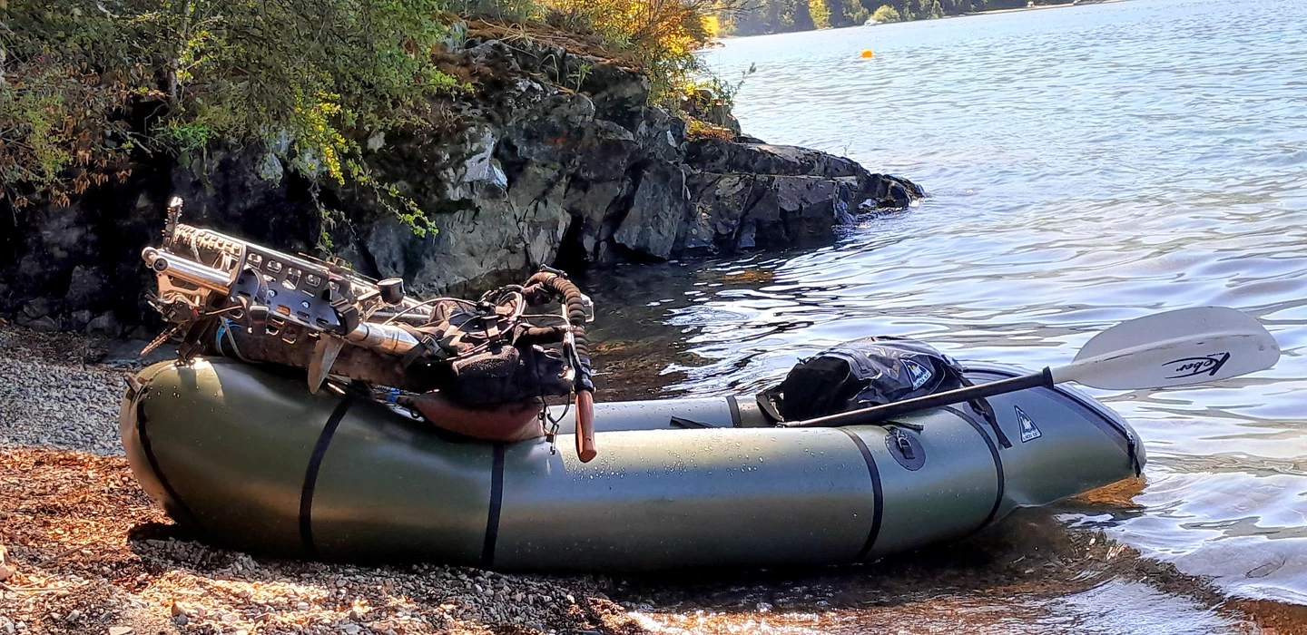 Olivgrünes Packraft mit Ausrüstung am Kiesufer, Felswand im Hintergrund, ruhiger See bei Tageslicht