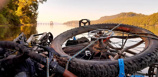 Detailaufnahme eines Mountainbike-Rads mit Scheibenbremse, im Hintergrund ein ruhiger See mit Anlegesteg und bewaldeten Ufern bei Sonnenaufgang