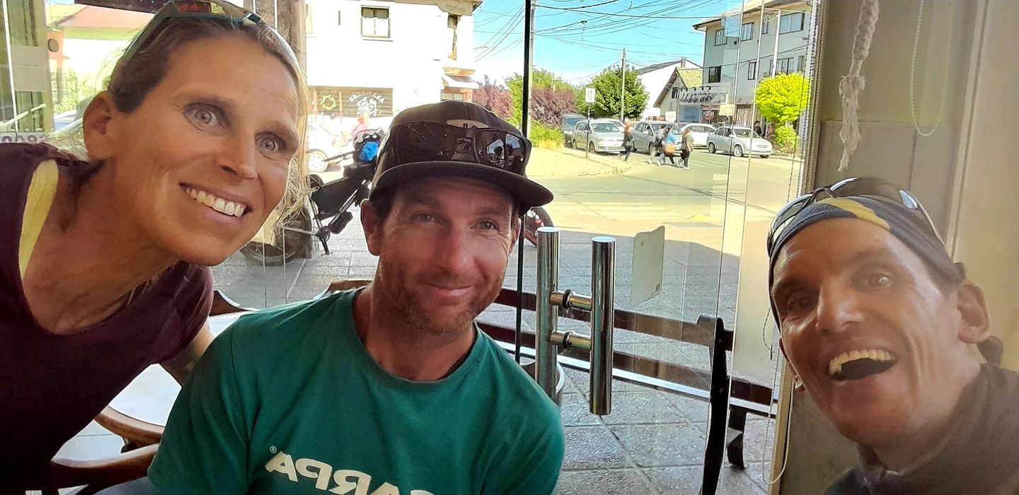 Drei lachende Personen in Radfahrer-Kleidung sitzen an einem Glastisch, Stadtstraße im Hintergrund