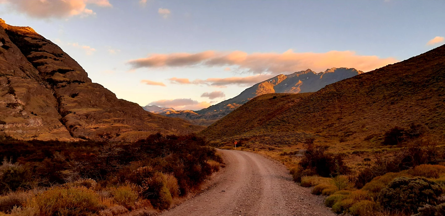 Gewundene unbefestigte Schotterpiste führt durch karges Bergland mit herbstlich gefärbter Vegetation und Bergketten im Hintergrund