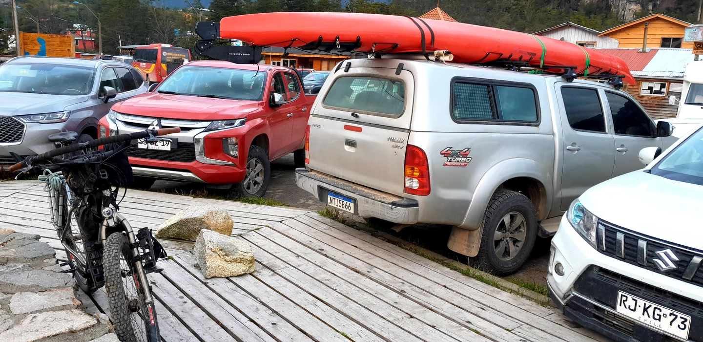 Silberner Pickup mit rotem Kanu auf dem Dach, daneben weitere Fahrzeuge auf Holzplattform mit Felsbrocken