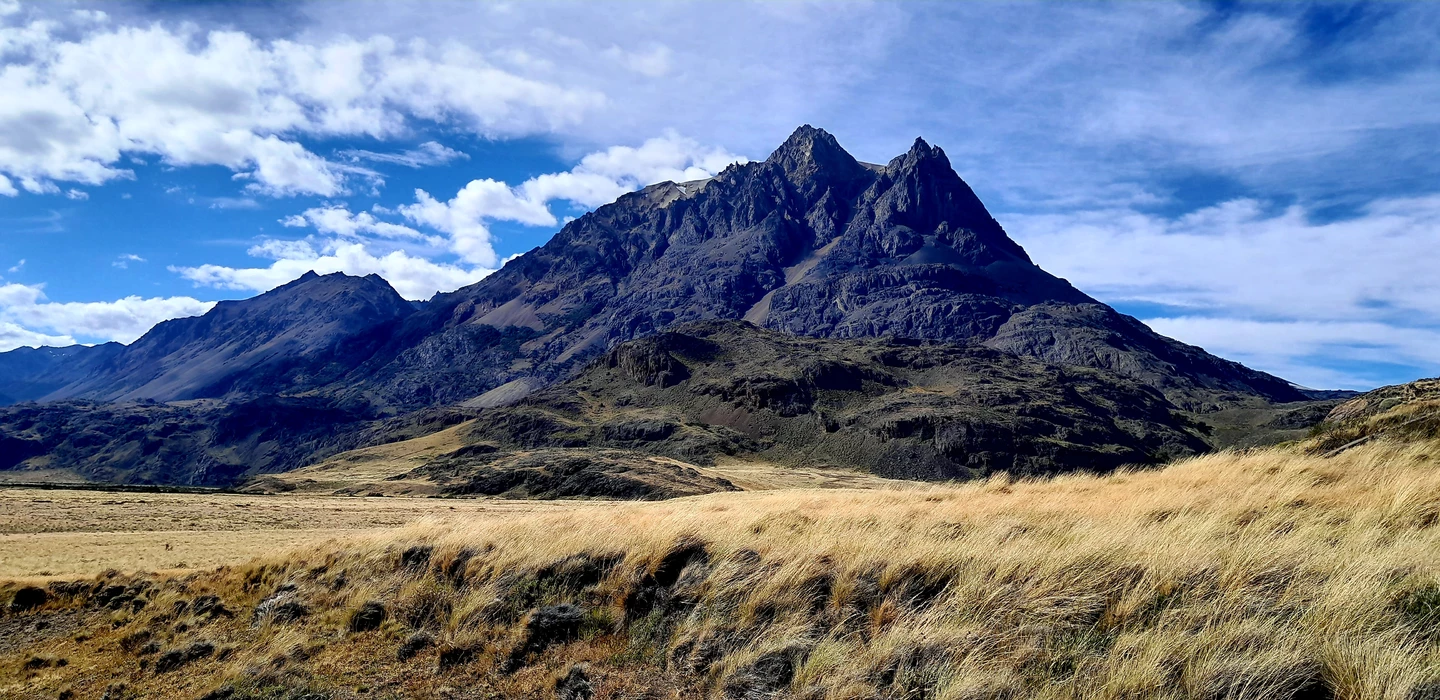 Steile, felsige Berggipfel in grau-blauer Färbung, umgeben von goldgelber Steppenvegetation und blauem Himmel mit Wolken