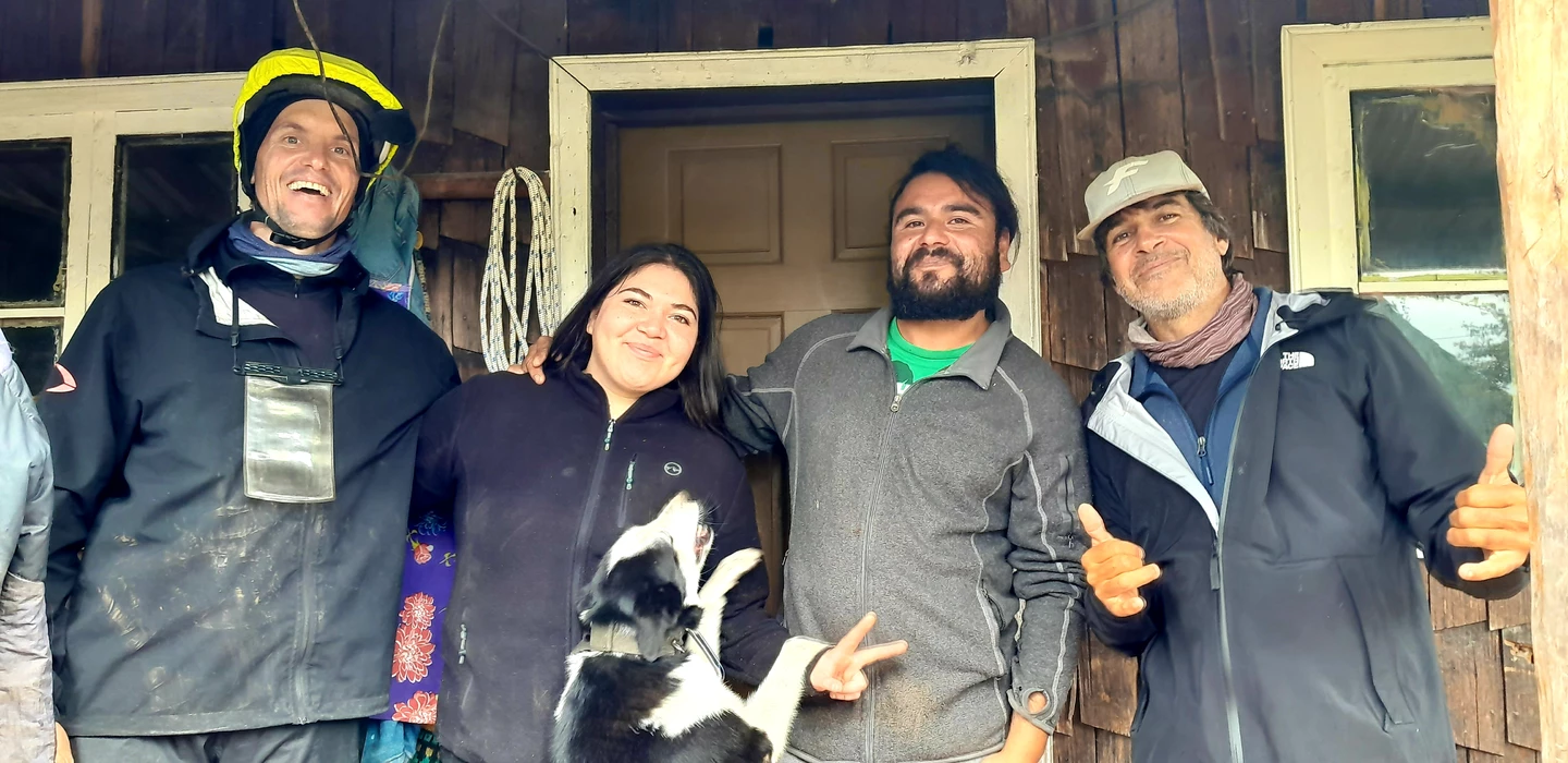 Vier Personen und ein schwarz-weißer Hund vor Holzhaus, in Outdoorkleidung, lächelnd