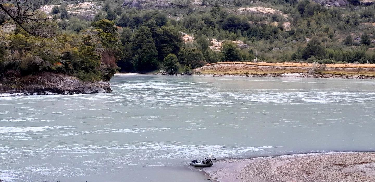 Türkisfarbener Fluss mit Packraft am Ufer, umgeben von dichten Wäldern und Felsen in patagonischer Landschaft