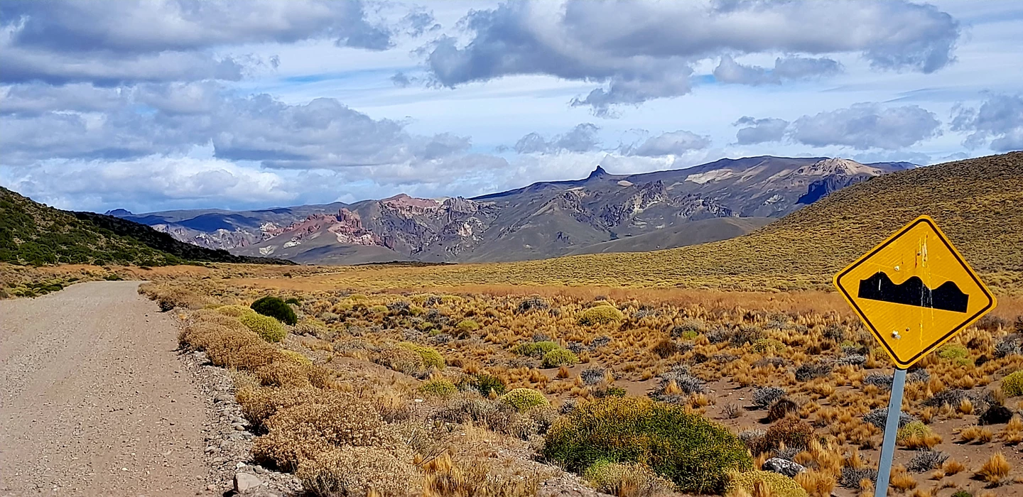 Weite Landschaft mit Schotterpiste, gelbe Grassteppen, markante Bergkette am Horizont und Warnschild für Straßenunebenheiten
