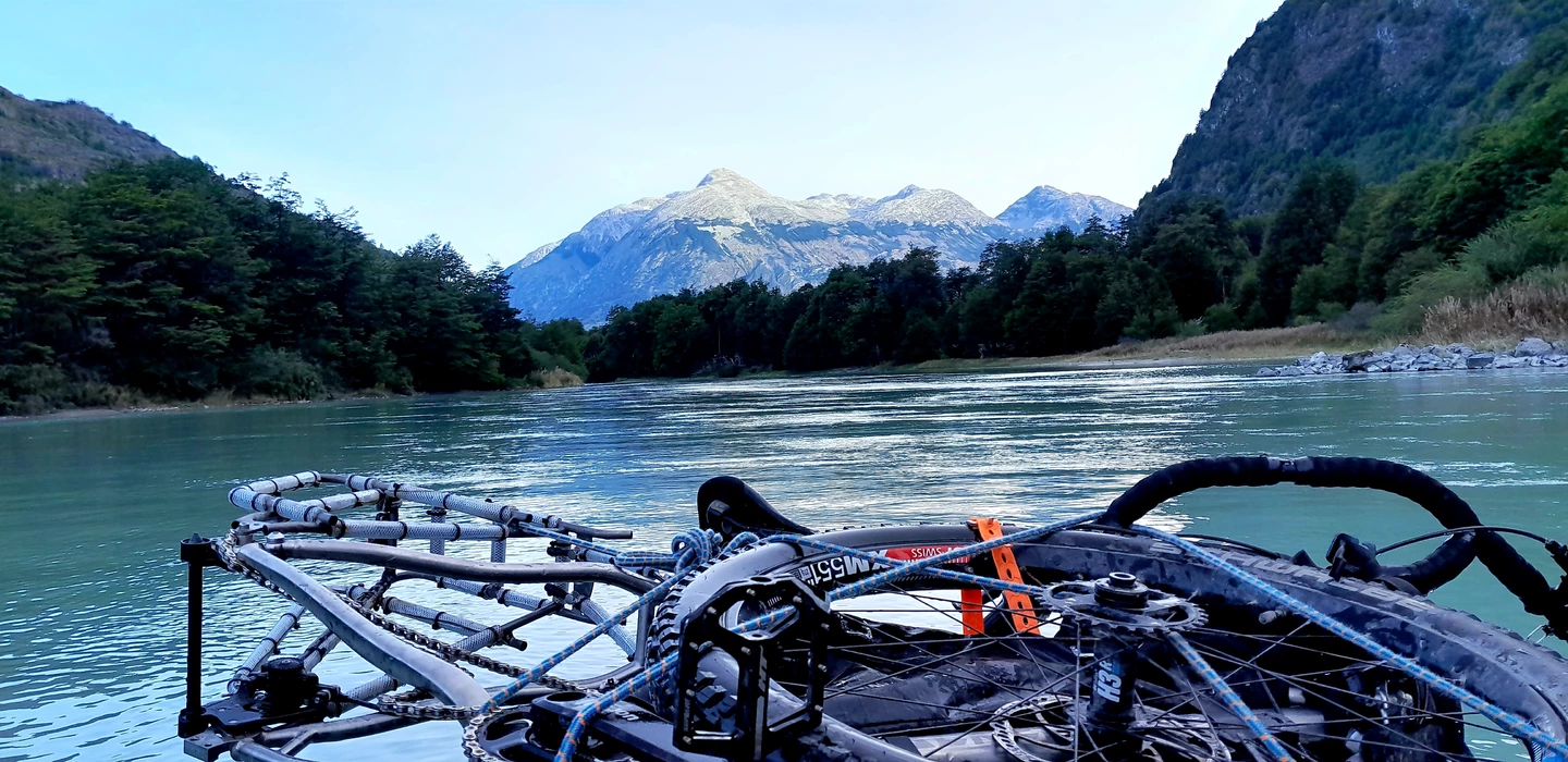 Fahrrad-Rahmen auf türkisgrünem Fluss, umrahmt von bewaldeten Bergen und schneebedeckten Gipfeln in Patagonien