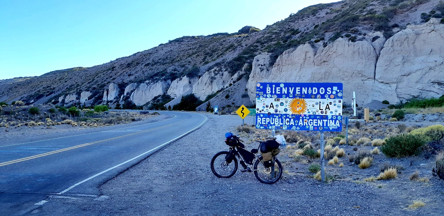 Fahrrad an Grenzschild der Argentinischen Republik, Gebirgslandschaft mit weißen Felsen und Bergstraße im Hintergrund