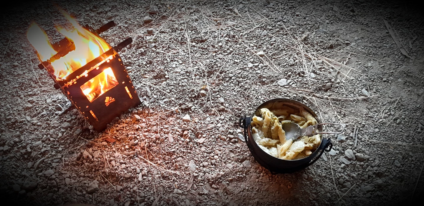Kleine Feuerstelle mit lodernden Flammen neben schwarzem Kochtopf auf steinigem Untergrund mit trockenen Gräsern
