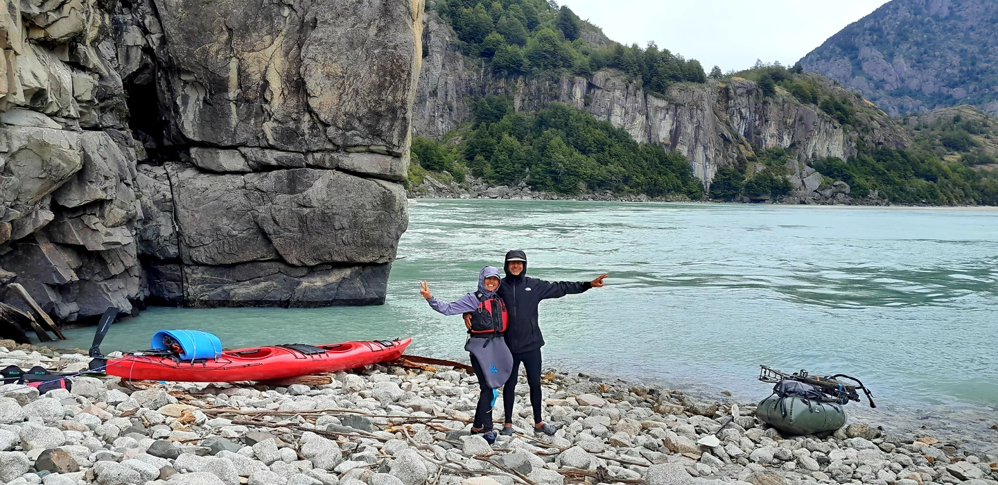 Zwei Kayak-Fahrer in Paddler-Ausrüstung an felsiger Küste mit rotem Kajak, Bergpanorama und türkisfarbenem Fluss im Hintergrund
