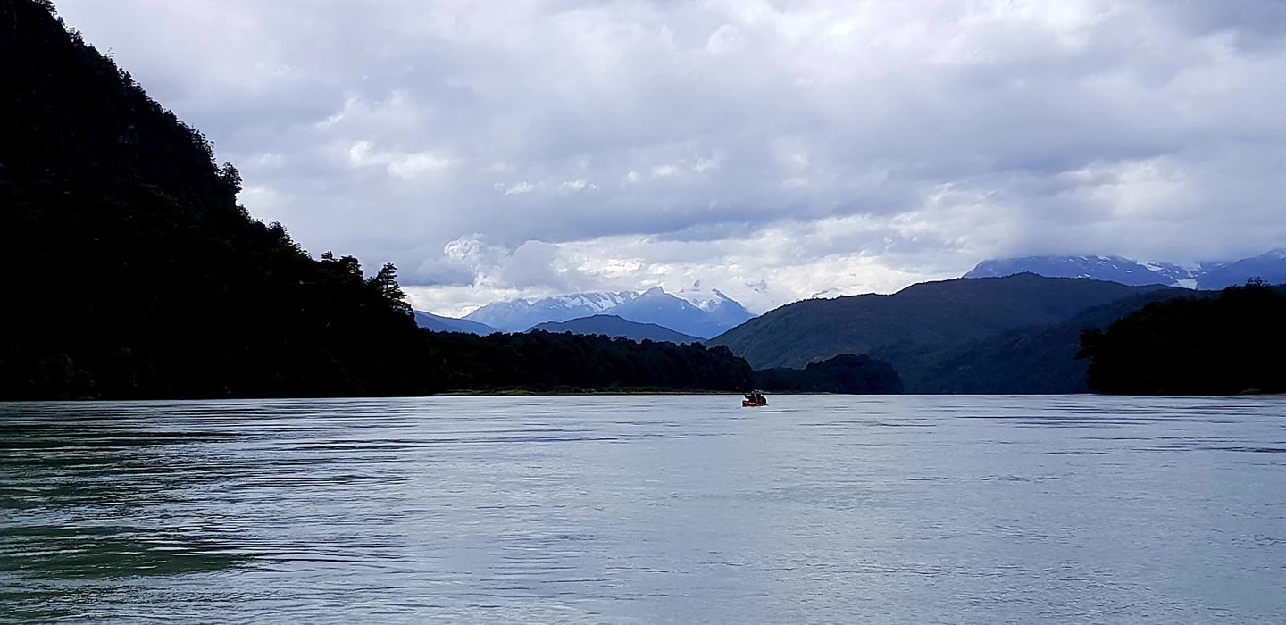 Weite Seelandschaft mit dunkelgrünen Wäldern, schneebedeckten Bergen und einem einsamen roten Kajak auf ruhigem Wasser