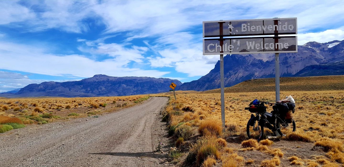 Unbefestigte Landstraße durch karges Grasland, im Hintergrund verschneite Berge, Willkommensschild und Fahrrad mit Gepäck