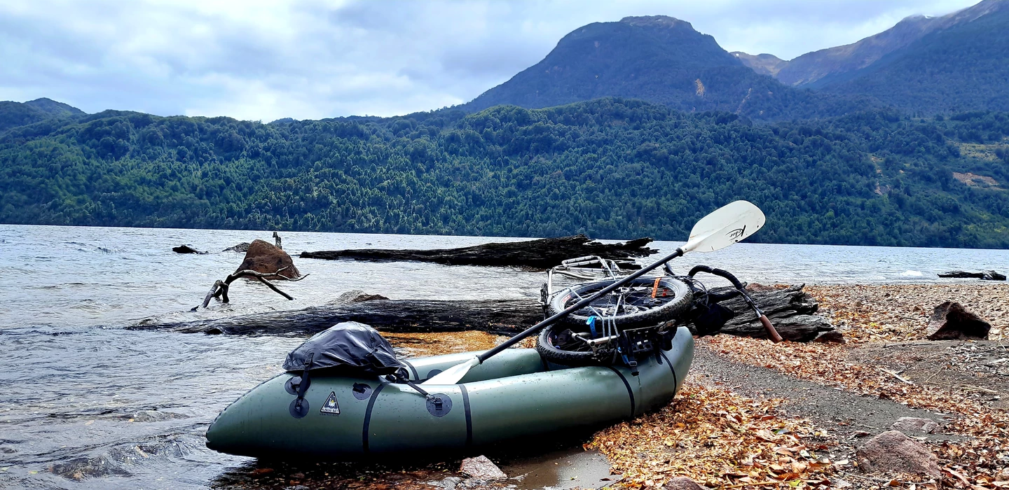 Grünes Packraft am Ufer eines Sees, umgeben von bewaldeten Bergen und Felsbrocken