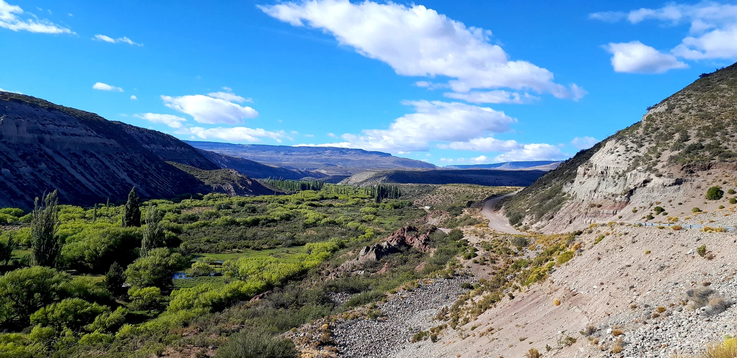 Weite Berglandschaft mit grünem Tal, Schotterpiste, felsigen Hängen und blauem Himmel mit Wolken in Patagonien