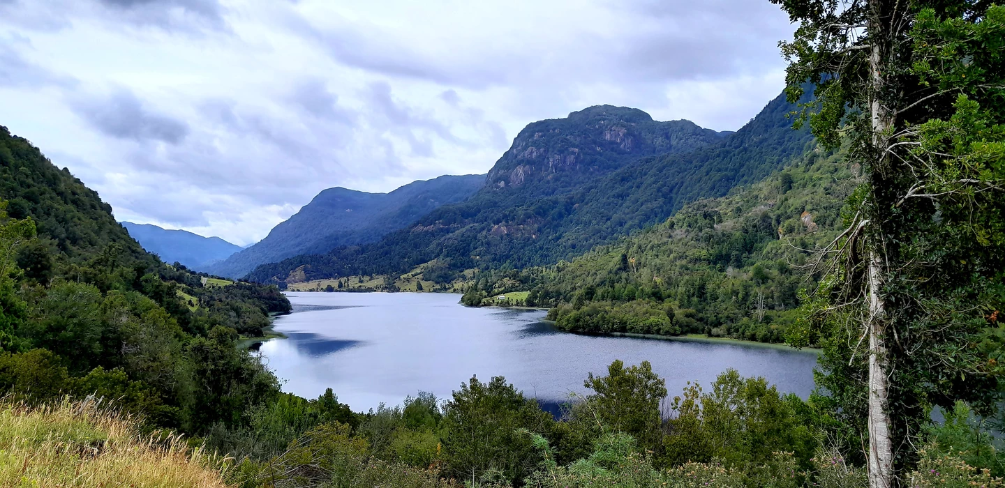 Weiter Bergsee umgeben von dichten Wäldern, steile Berge im Hintergrund, bewölkter Himmel, saftig grüne Landschaft