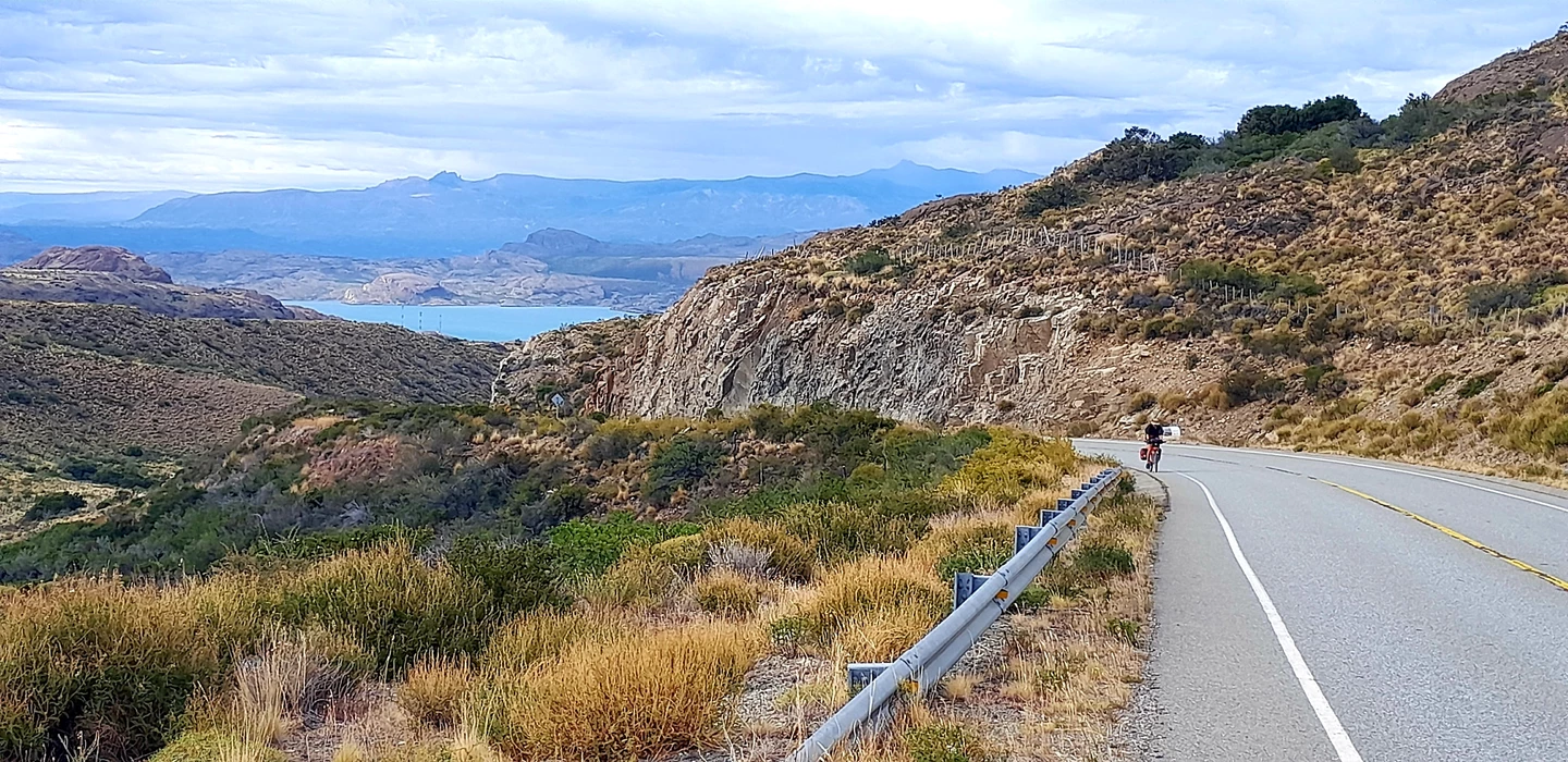 Kurvige Asphaltstraße durch felsige Berglandschaft, ein Radfahrer fährt bergauf, blauer See im Hintergrund, weite Bergketten
