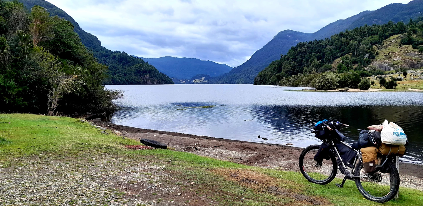Beladenes Fahrrad am Ufer eines spiegelglatten Sees, umgeben von bewaldeten Bergen und weiter Landschaft