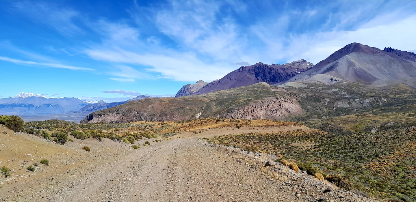 Unbefestigte Schotterpiste durch karges Bergland mit vulkanischen Gipfeln, weitem Himmel und Vegetation in Braun- und Grüntönen