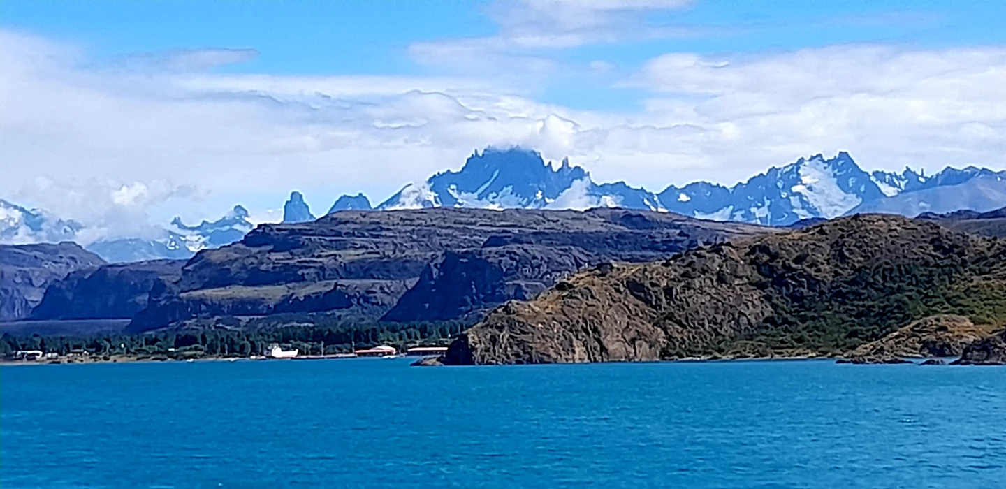 Türkisblaue Wasserfläche mit felsiger Küste, dahinter schneebedeckte Bergketten der Patagonischen Anden in blau-weißen Tönen