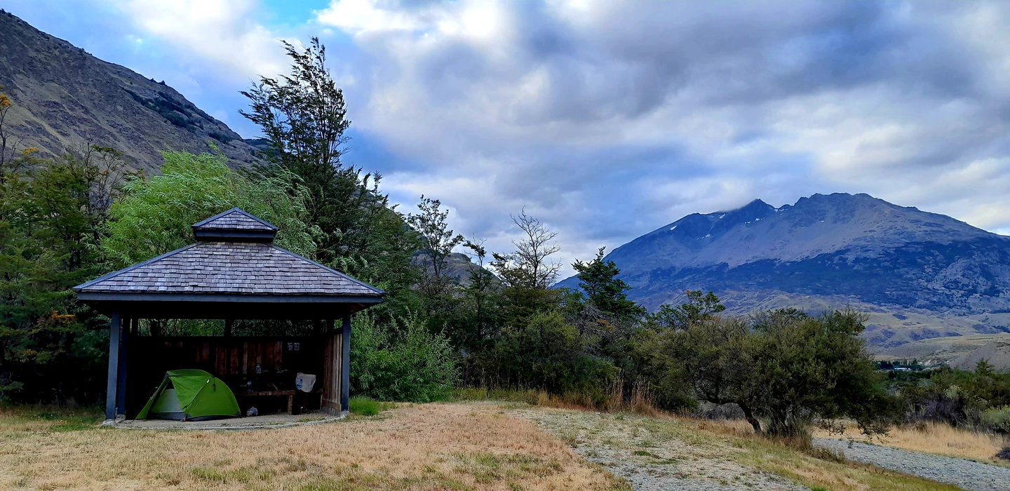 Grünes Zelt unter einem Holzpavillon, umgeben von Bäumen und Berglandschaft in Patagonien