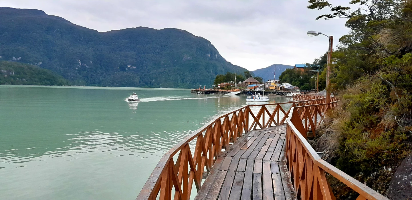 Holzsteg mit Geländer führt an türkisgrünem Bergsee vorbei, Boote am Steg, bewaldete Berge im Hintergrund