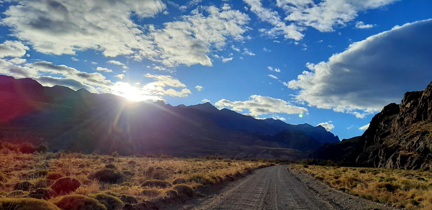 Unbefestigte Straße durch trockene Graslandschaft, im Hintergrund Berge mit Sonnenlicht und dramatischem Wolkenhimmel