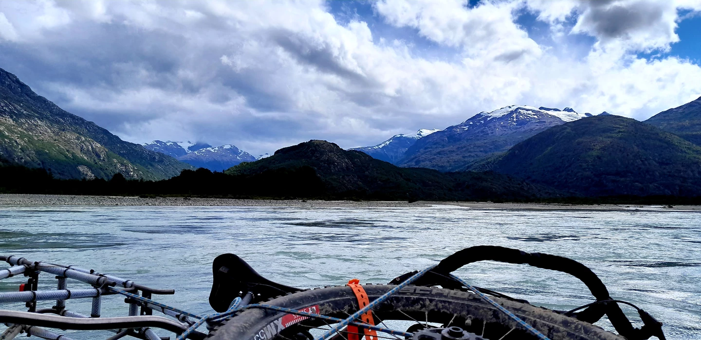 Fahrradlenker im Vordergrund, dahinter weiter Fluss und schneebedeckte Berge der Patagonischen Anden
