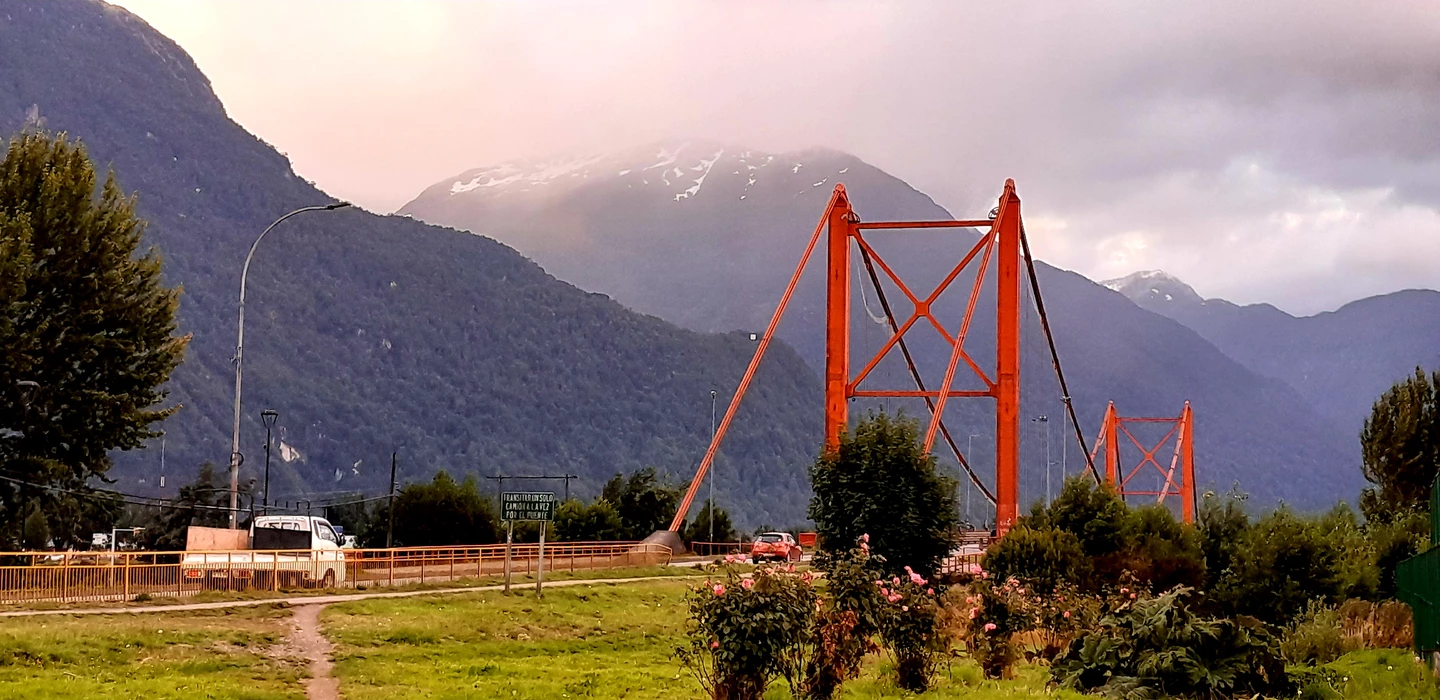 Orangerote Hängebrücke vor schneebedeckten Bergen, grüne Wiese im Vordergrund, Bäume und Sträucher