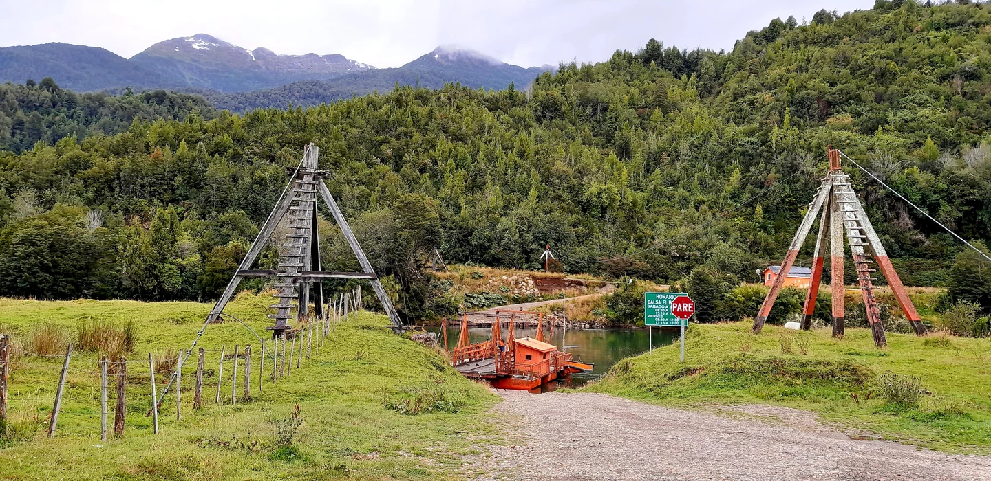 Alte Holzpylone einer Seilfähre an einem Fluss, grüne Wiese, Bergwald im Hintergrund, Patagonische Landschaft