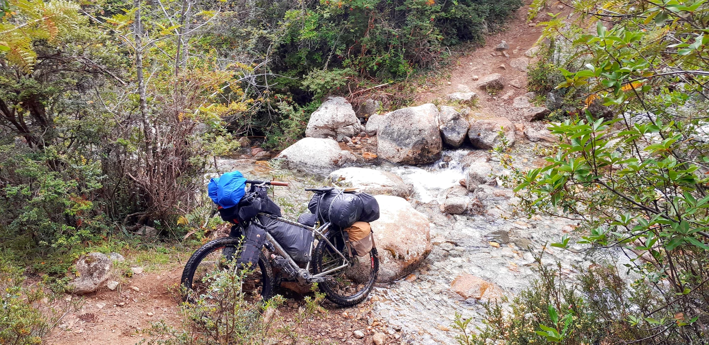 Voll bepacktes Mountainbike lehnt an Felsen neben einem steinigen Gebirgsbach, umgeben von dichter Vegetation