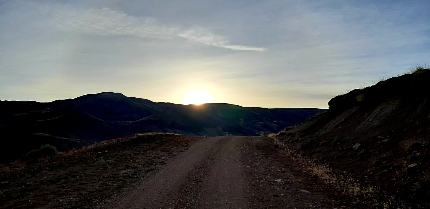 Unbefestigte Bergstraße bei Sonnenaufgang, Schotter und Geröll, Berge im Hintergrund, Morgenlicht