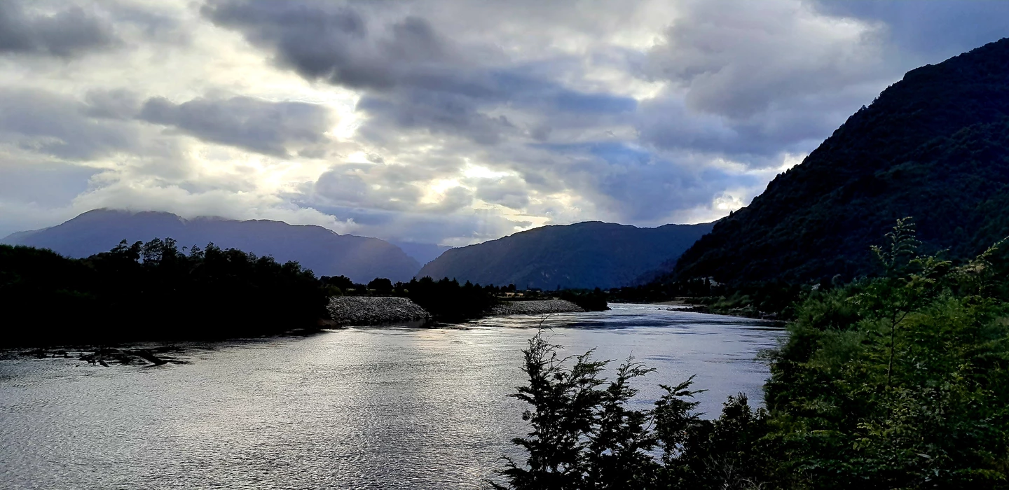 Breiter Fluss mäandriert durch grüne Täler, umgeben von dunklen Bergen unter dramatischem Wolkenhimmel