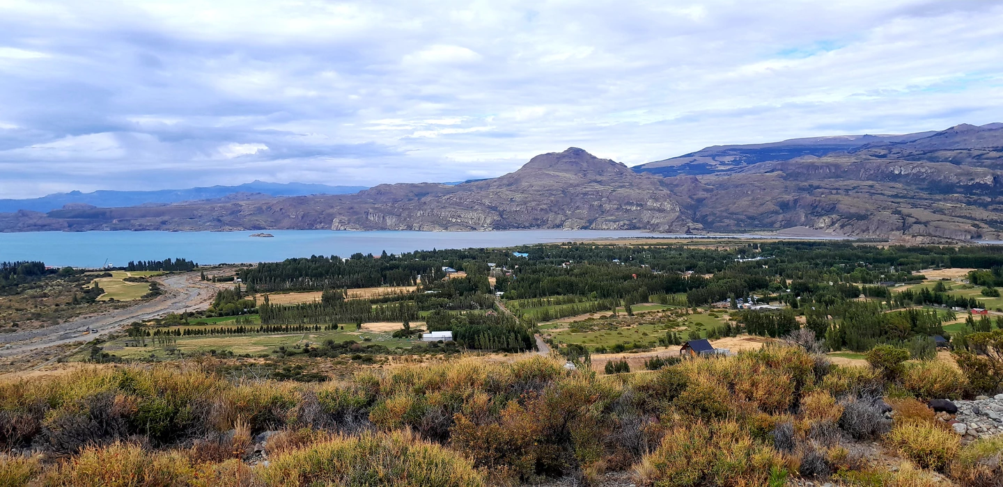 Weite Patagonien-Landschaft mit türkisfarbenem See, grünem Tal, Nadelwäldern und kargen Bergketten im Hintergrund