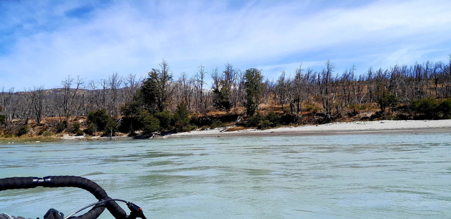Blick vom Wasser auf felsiges Ufer mit kahlen Bäumen, türkisfarbener Fluss, Bergkulisse im Hintergrund