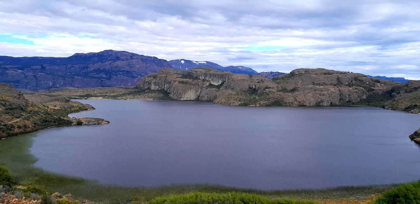 Weiter blaugrauer See umgeben von felsigen Hügeln, Bergkulisse mit schneebedeckten Gipfeln im Hintergrund, bewölkter Himmel