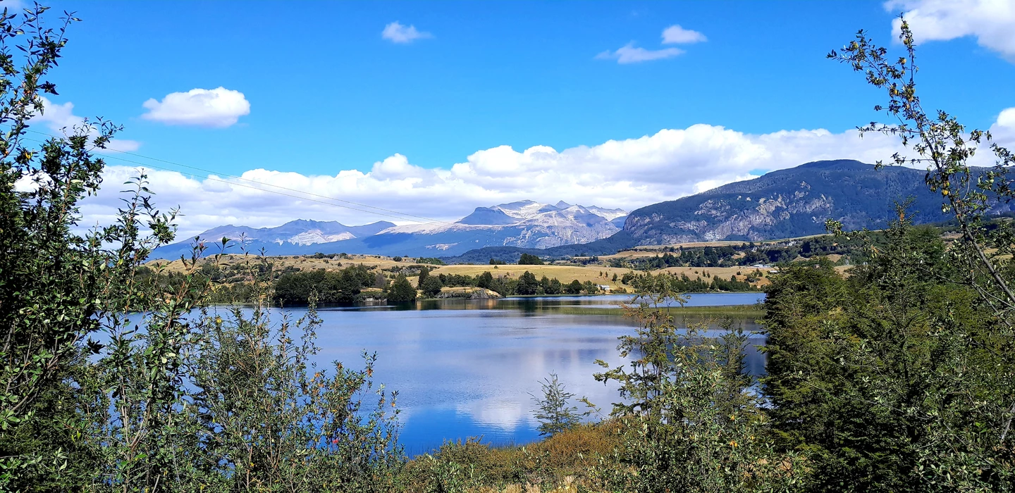 Blick auf einen ruhigen Bergsee, umgeben von Bäumen, mit schneebedeckten Bergketten im Hintergrund unter blauem Himmel