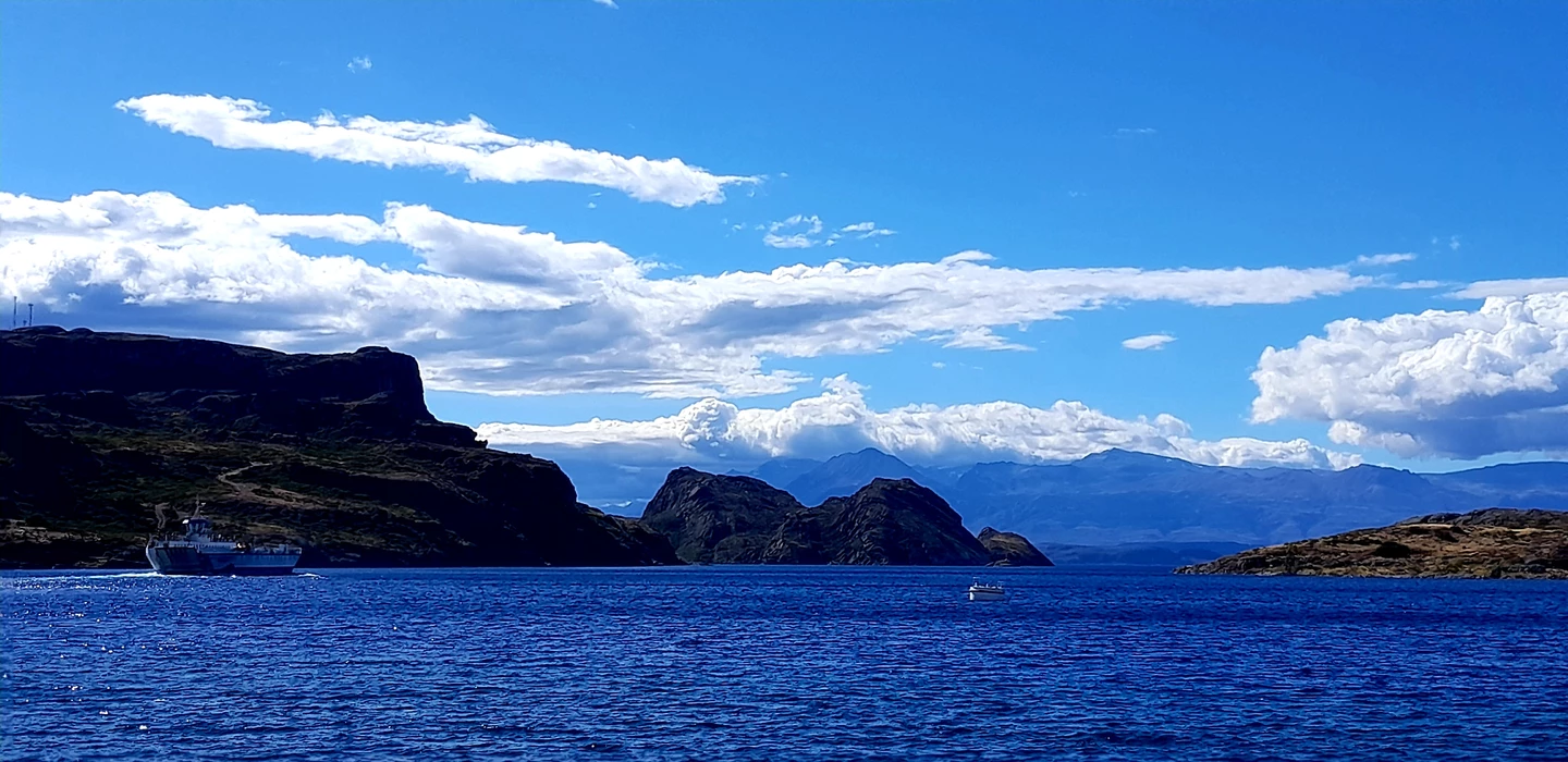 Blick auf blaues Gewässer mit Bergpanorama, weißen Wolken und einem Schiff vor felsigen Ufern in Patagonien