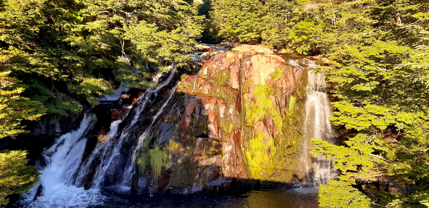 Szenischer Wasserfall mit rötlich-braunen Felsen, umgeben von grünen Bäumen, Wasser fließt über gestaffelte Felsstufen