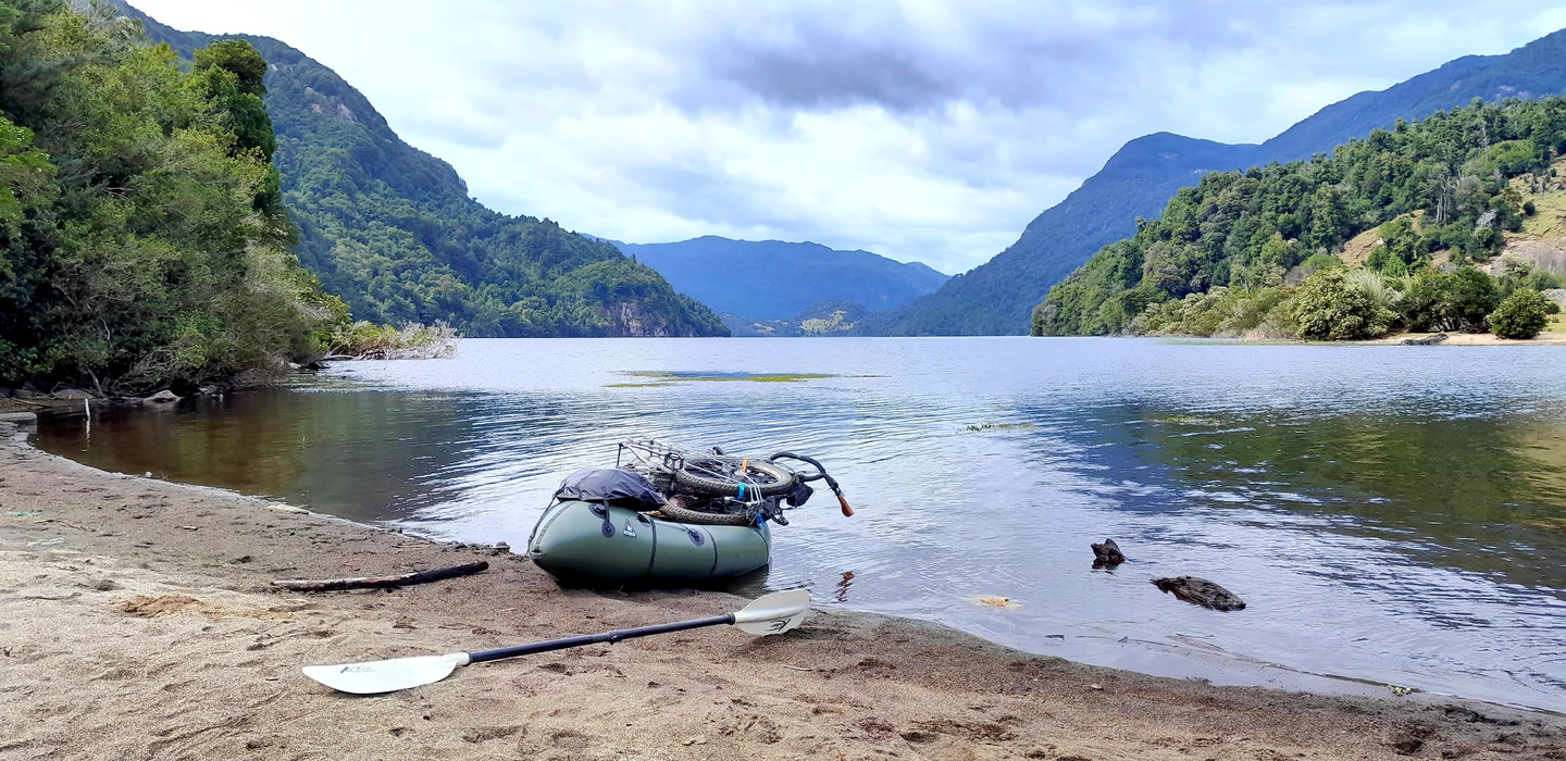 Grünes Packraft mit Fahrrad am sandigen Ufer eines spiegelglatten Bergsees, umgeben von dicht bewaldeten Berghängen