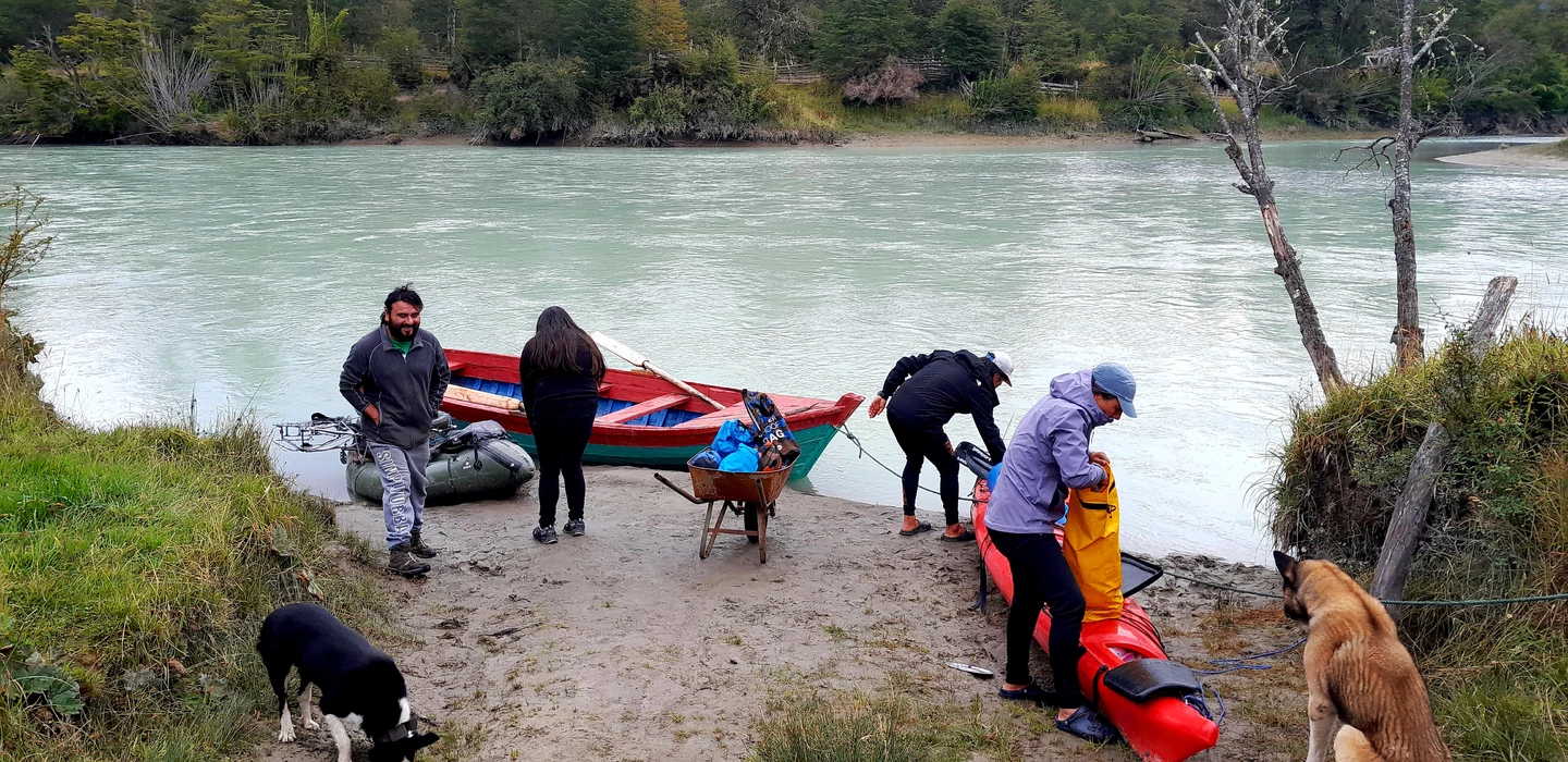 Vier Personen und zwei Hunde bereiten Boote und Ausrüstung an einem türkisfarbenen Fluss mit Wald im Hintergrund vor