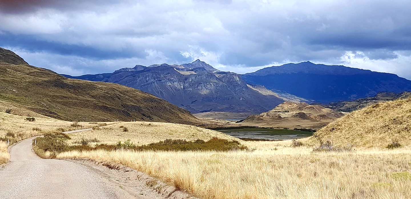 Weite Graslandschaft mit kurvigem Schotterweg, im Hintergrund mächtige blaue Berge unter dramatischem Wolkenhimmel