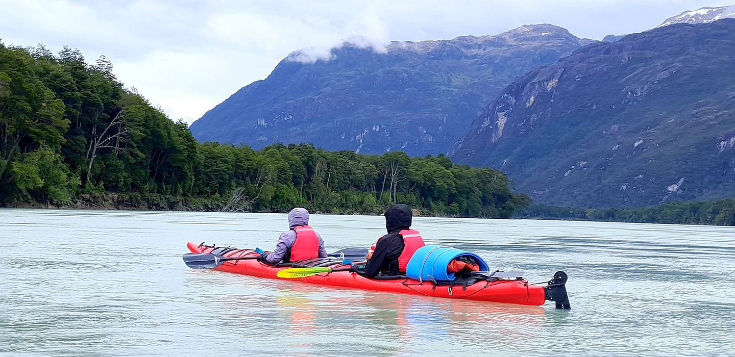 Zwei Kayakfahrer in roten Booten auf einem glatten See, umgeben von grünen Wäldern und blauen Berggipfeln mit Wolken