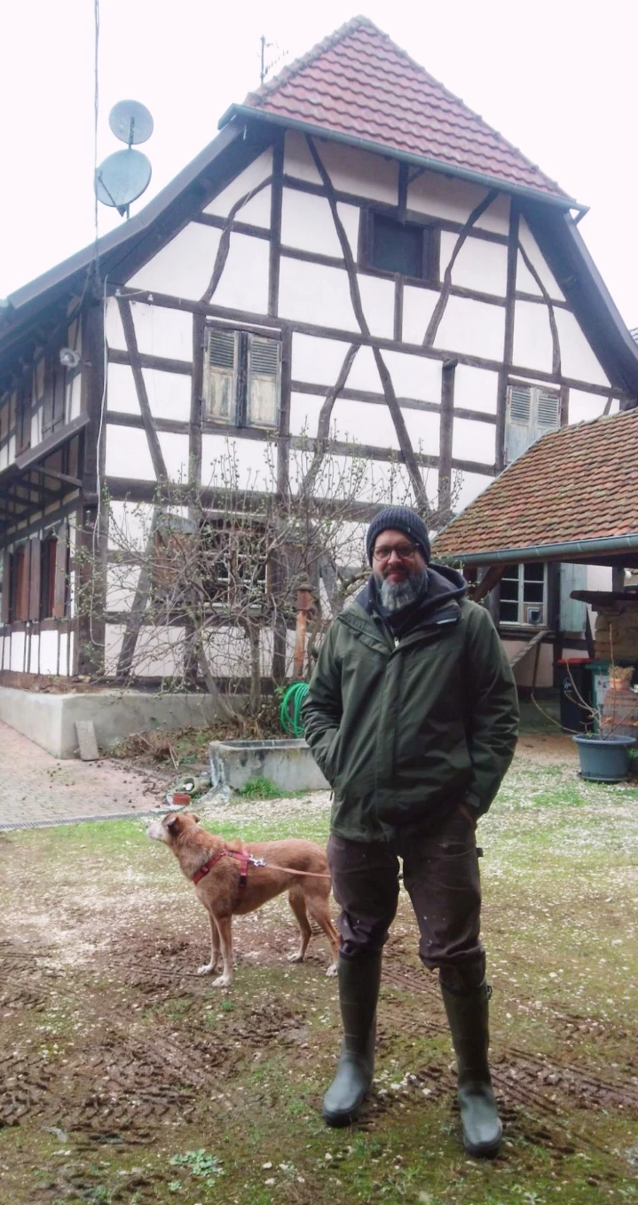 Älterer Mann mit Bart und grüner Jacke steht mit braunem Hund vor einem weißen Fachwerkhaus mit Satellitenschüssel