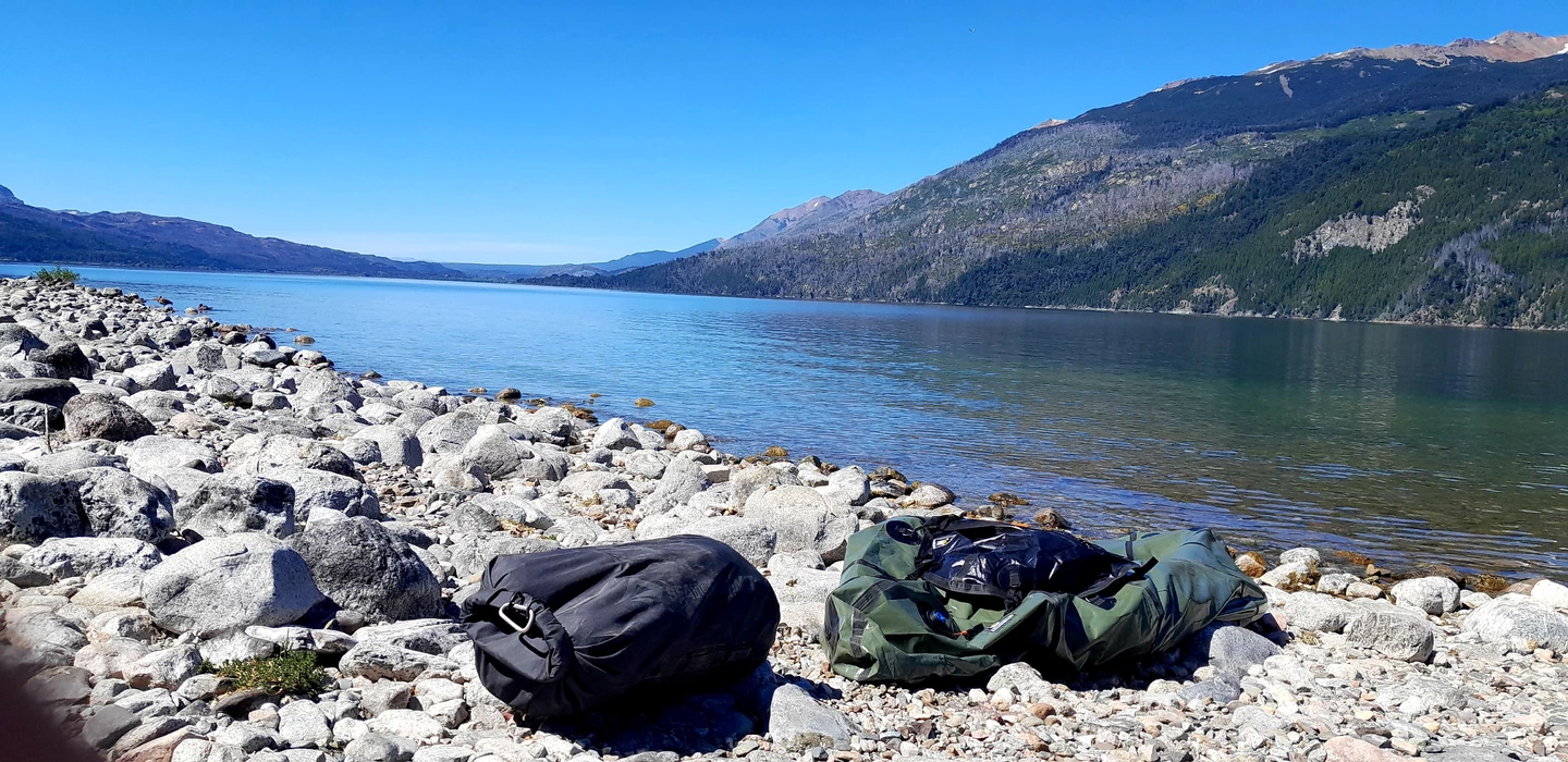 Weite Berglandschaft mit blauem See, Felsufer und zwei Wanderrucksäcken im Vordergrund