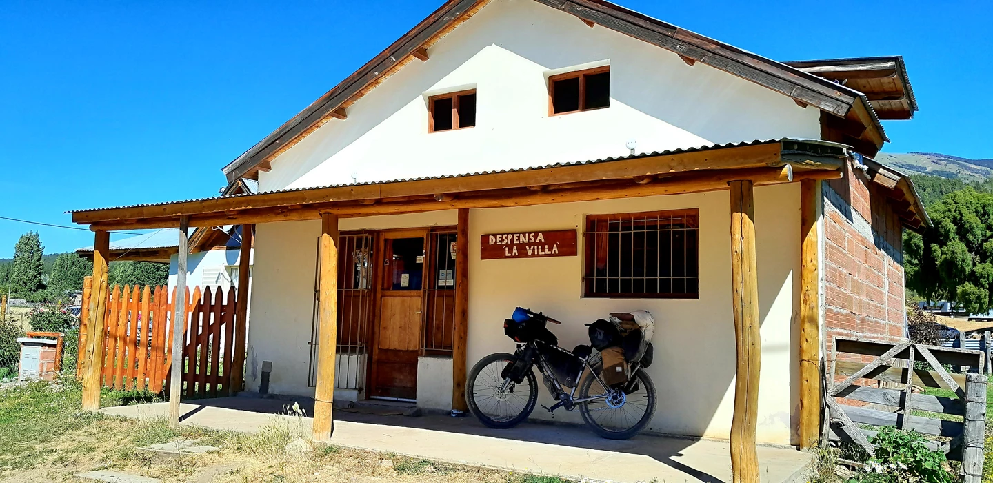 Weißes Backsteinhaus mit Holzveranda, Fahrrad mit Packtaschen, Ortsschild 'Despensa La Villa', Berglandschaft im Hintergrund