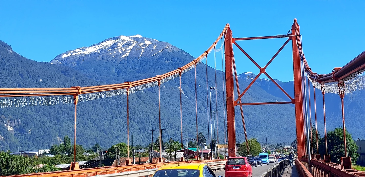 Rote Hängebrücke über Straße, dahinter schneebedeckte Berge und blauer Himmel, Autos fahrenen