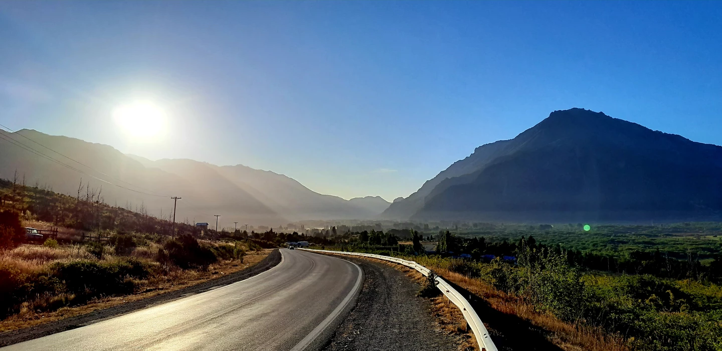 Sonnendurchflutete Landstraße mit scharfer Kurve, umgeben von herbstlichen Büschen und mächtigen Bergen im Hintergrund