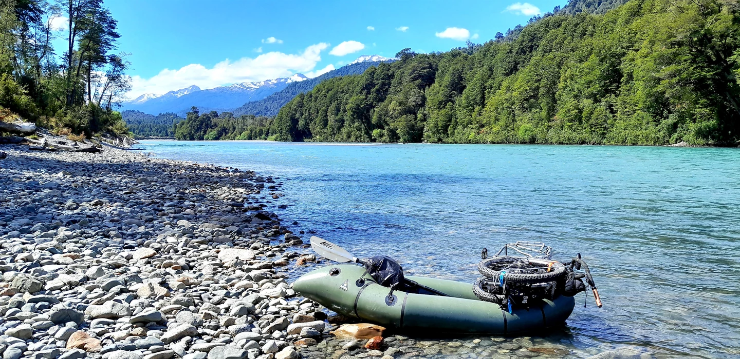 Grünes Packraft mit Laufrad auf Kieselstrand, dahinter türkisfarbener See, dicht bewaldete Berghänge und schneebedeckte Gipfel