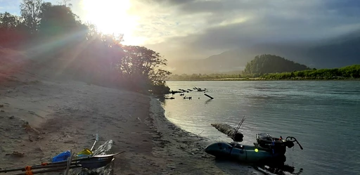 Zwei Packrafts am Ufer eines Flusses, Morgenlicht durchbricht Bäume, Berge im Hintergrund, nebliger Fluss