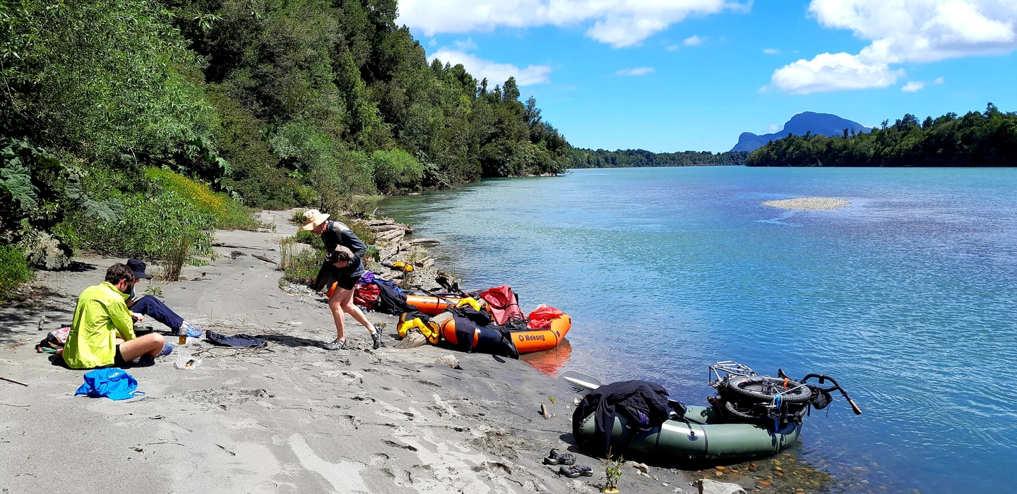 Zwei Packraft-Abenteurer an sandiger Flussufer mit Booten, grünem Wald und blauem Bergsee im Hintergrund