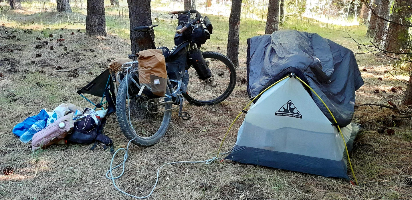 Mountainbike mit Seitentaschen neben grauem Zelt zwischen Kiefernstämmen, Campingausrüstung verstreut auf Nadelboden